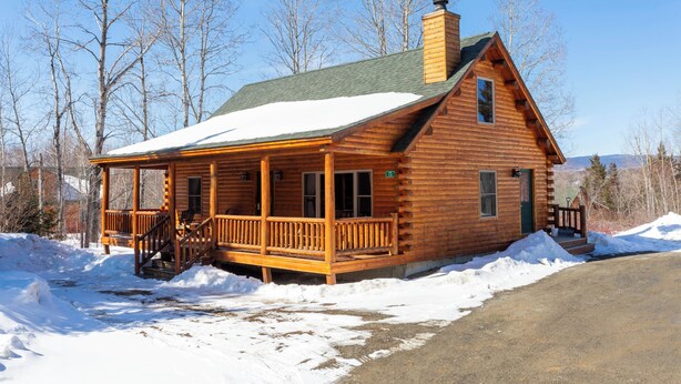A log cabin with a porch, a chimney, and a snow-covered roof.