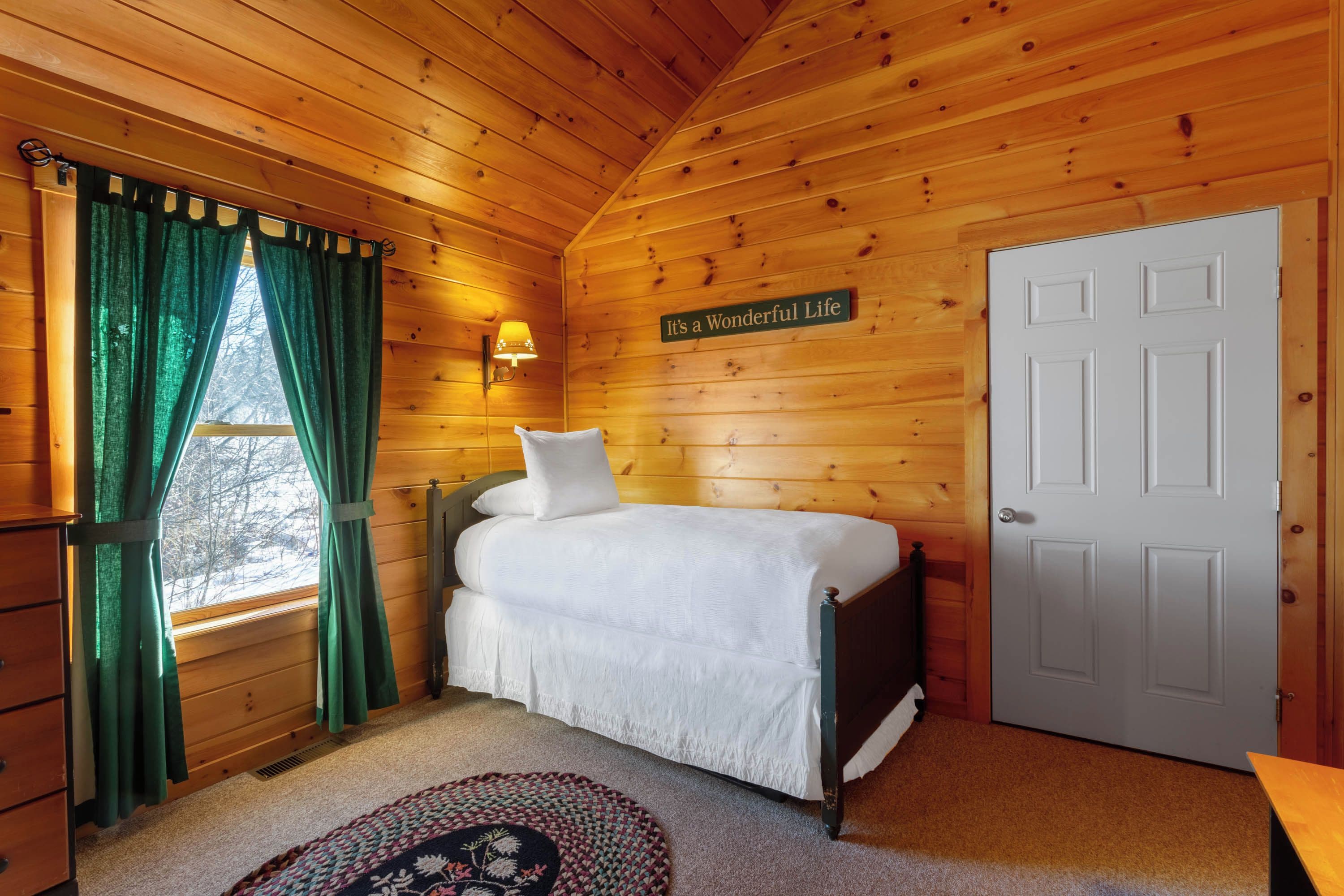 A wooden cabin bedroom with a bed, a wall-mounted lamp, and a door.