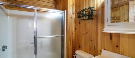 Ett badrum med träpanel på väggarna, en duschkabin med glasväggar, en toalett och ett handfat med en spegel ovanför.