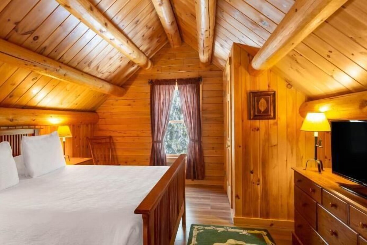 A wooden cabin bedroom with a bed, a TV, and a window with curtains.