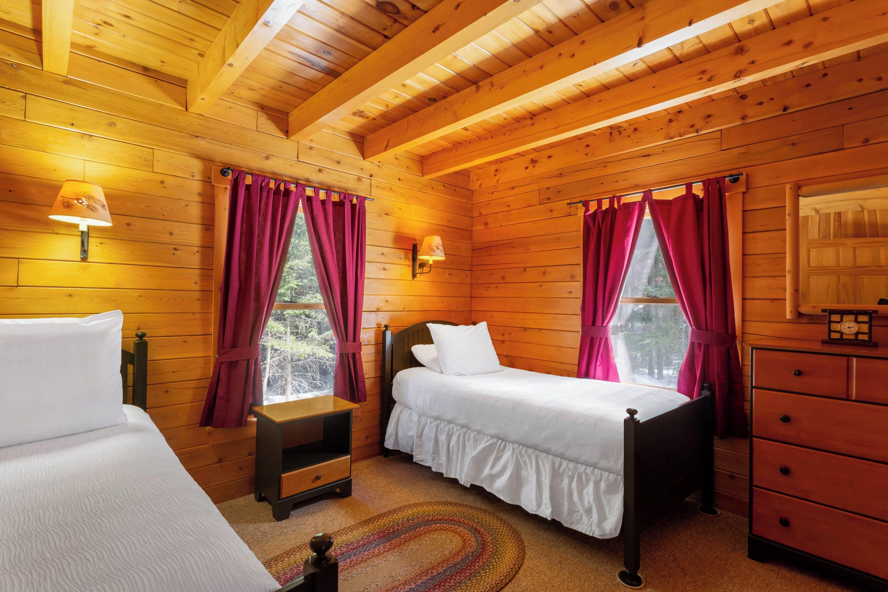A wooden cabin room with two beds, a nightstand, a dresser, and a window with curtains.