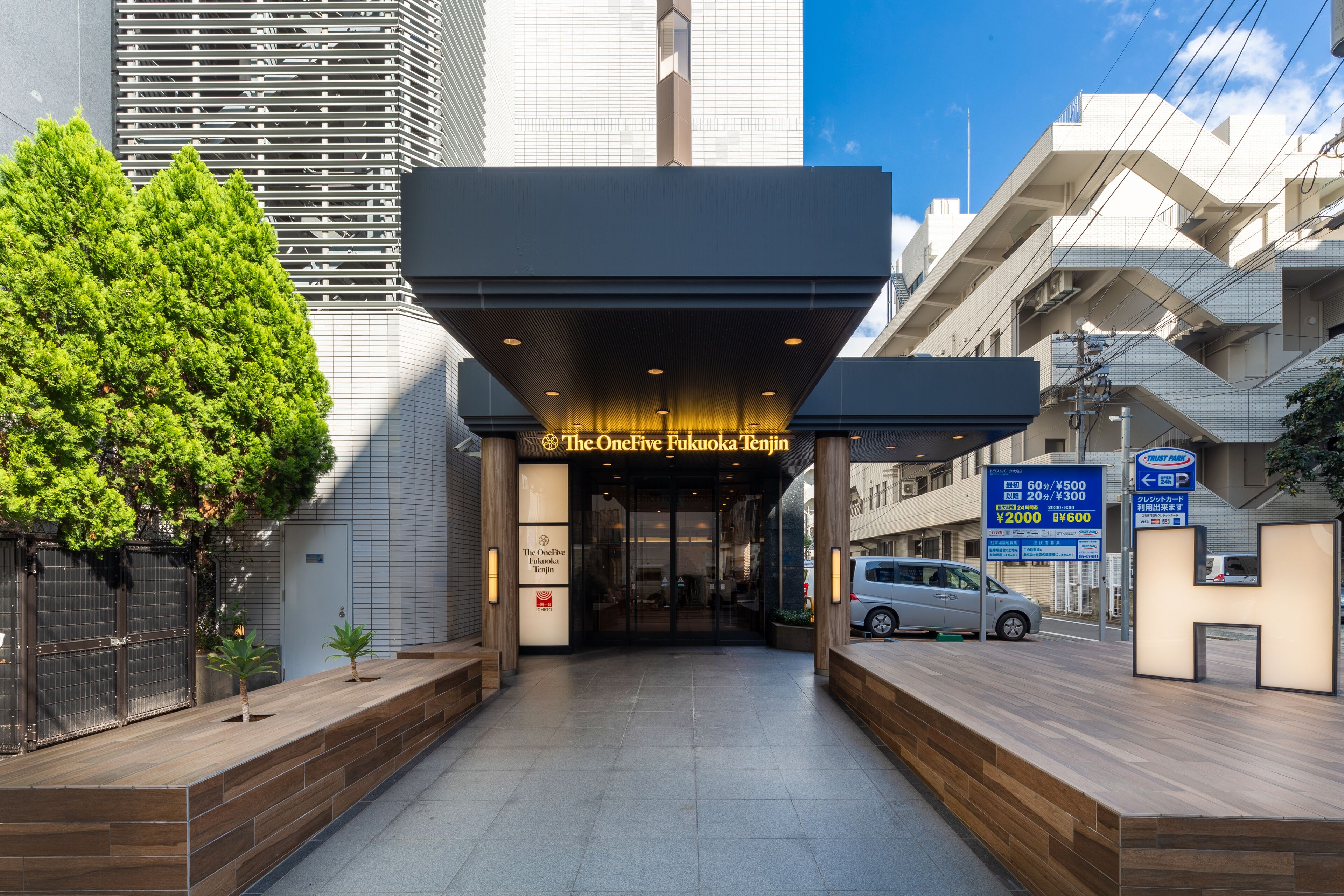 The entrance to The One Five Fukuoka Tenjin hotel with a modern design, a clear sky, and a parking sign visible.