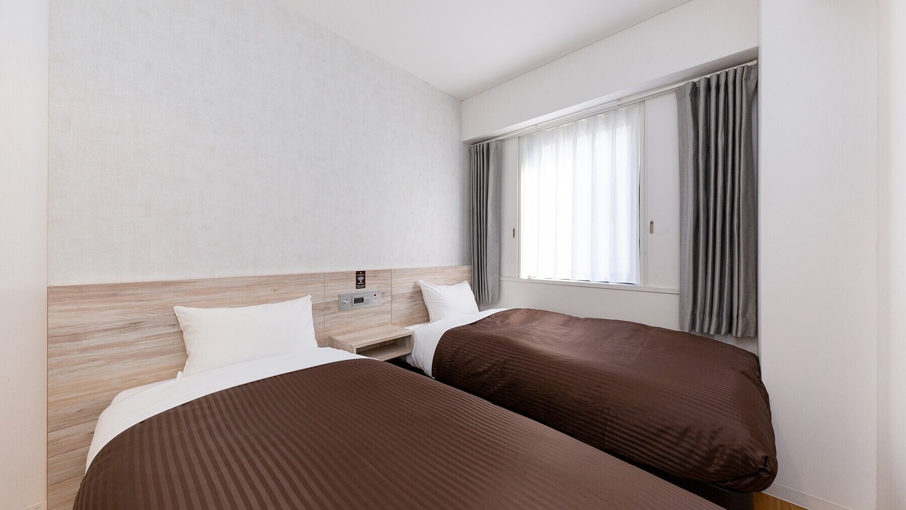 Ein Hotelzimmer mit zwei Betten, einem Holzkopfteil, einem Fenster mit Vorhängen und einer Bedienkonsole an der Wand.