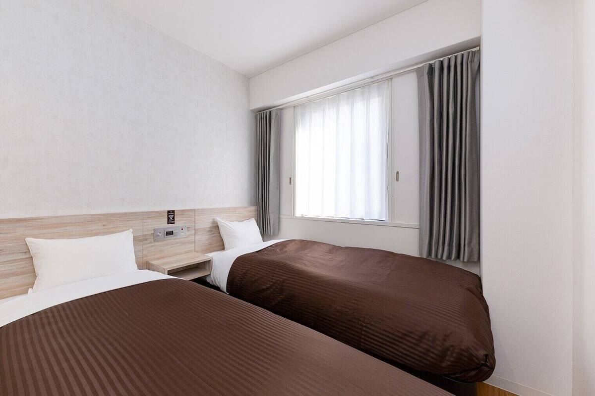 A hotel room with two beds, a wooden headboard, a window with curtains, and a control panel on the wall.