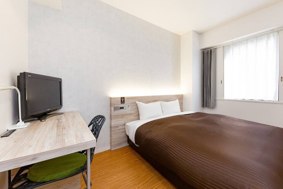 A hotel room with a bed, a desk with a television, a chair, and a window with curtains.