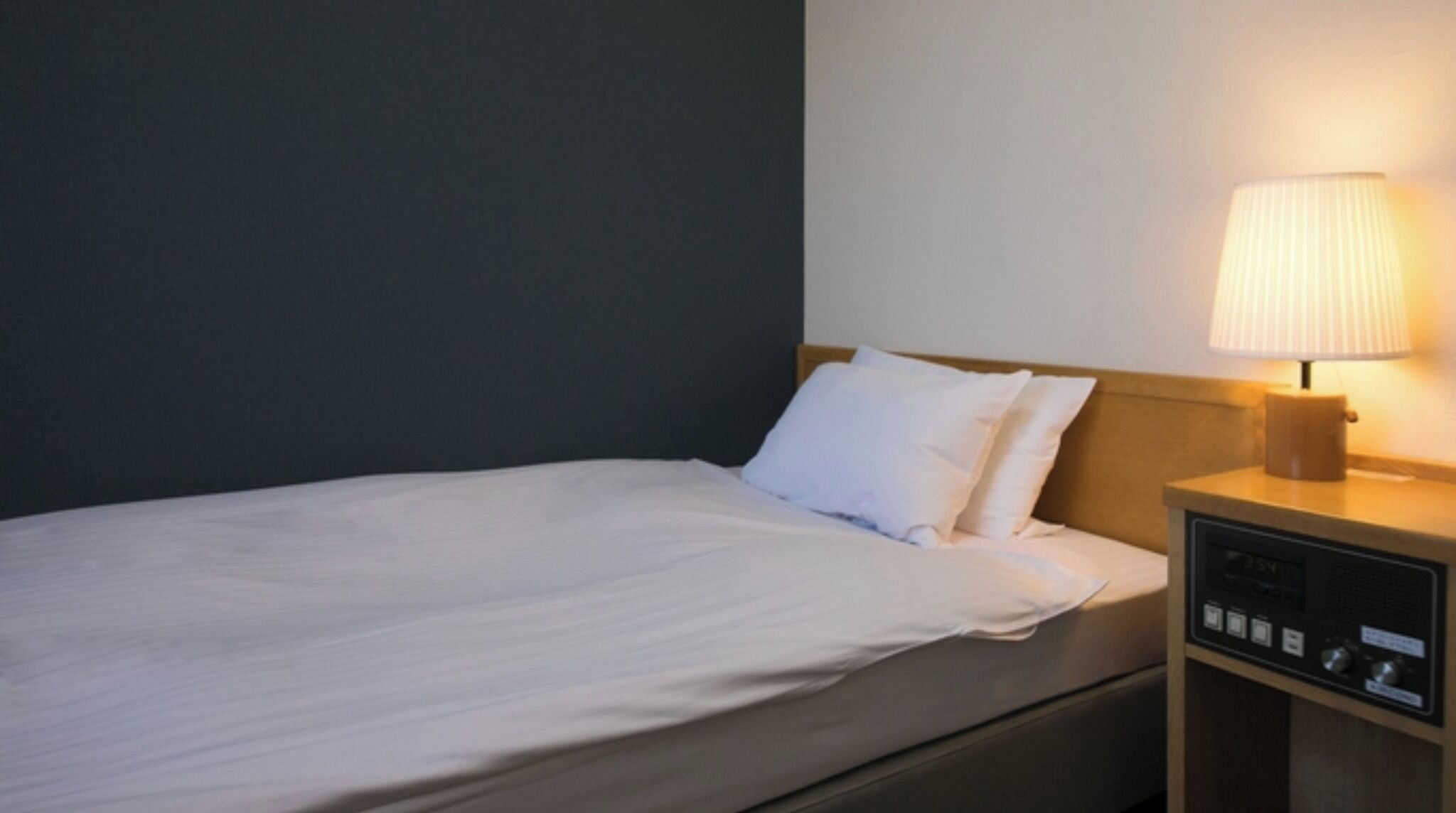 Una cama individual con ropa blanca, una mesita de noche con lámpara y un panel de control.