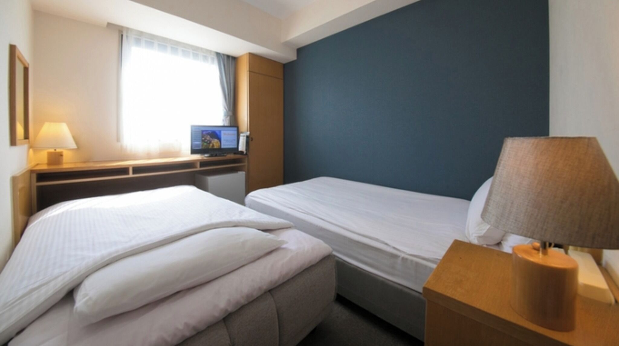 Una habitación de hotel con dos camas, un escritorio con computadora, una lámpara y una ventana con cortinas.