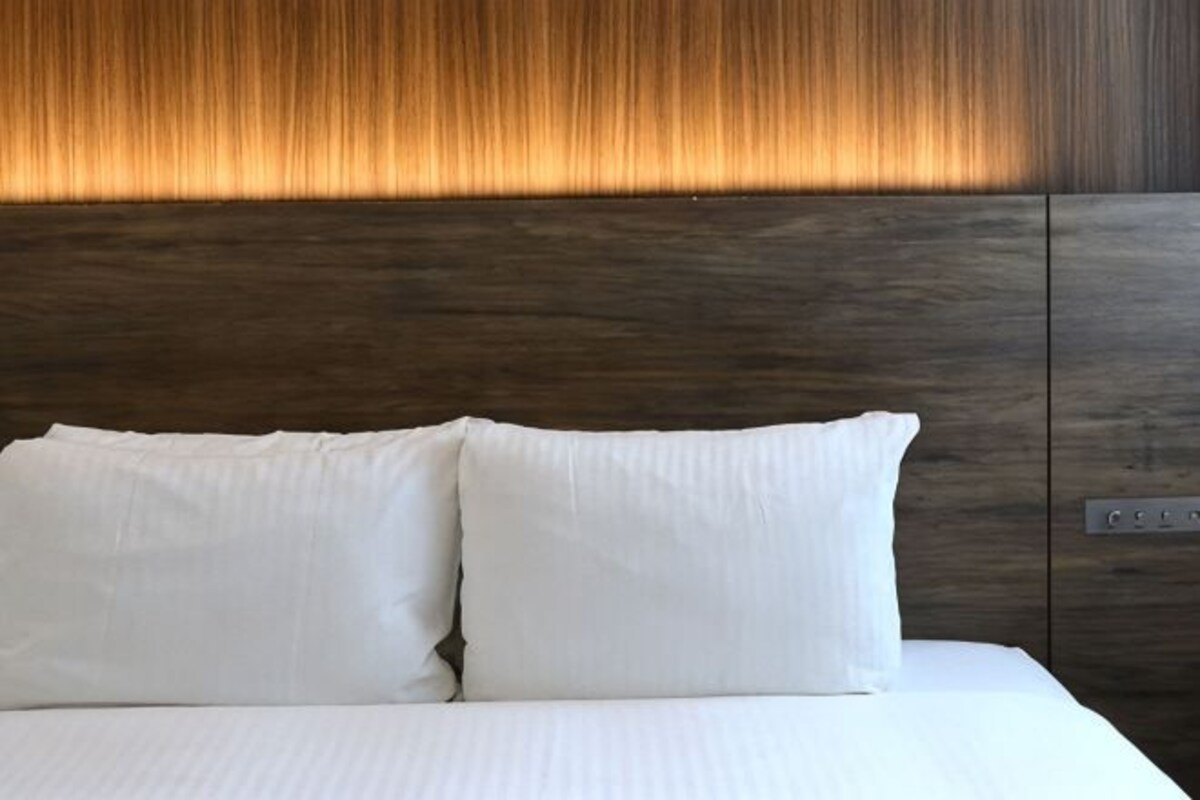 Una cama con ropa de cama y almohadas blancas, cabecera de madera con un panel iluminado y panel de control lateral.