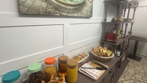 Un bufé de desayuno con bollería, café y zumo, una pintura de una escena callejera europea y una estantería de madera con objetos decorativos.