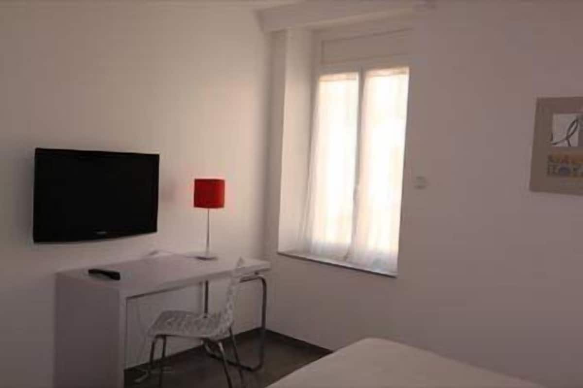 Una habitación pequeña y sencilla con un escritorio blanco, una silla, un televisor y una ventana con cortinas.