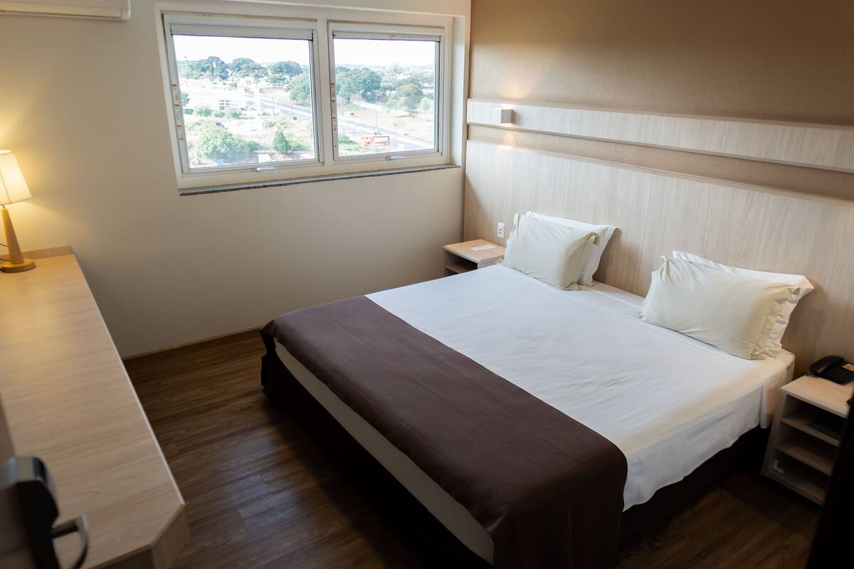 Habitación de hotel con una cama grande, una mesita de noche, una lámpara y una ventana con vistas al exterior.