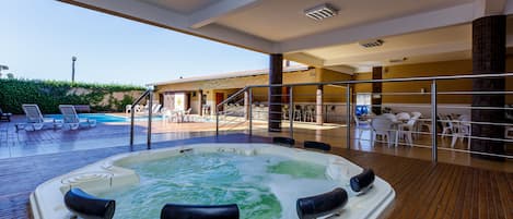 A hot tub on a wooden deck with a pool and lounge chairs in the background.