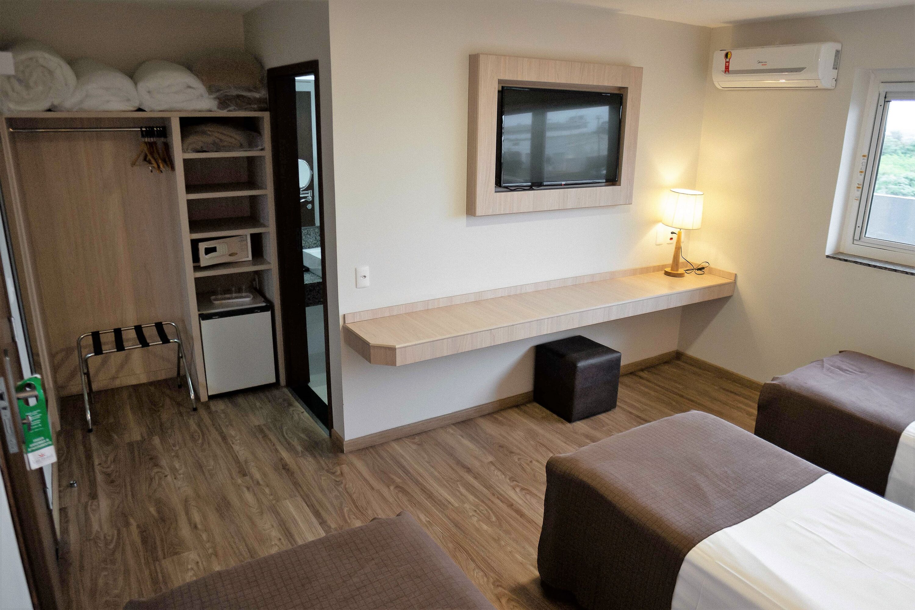Una habitación de hotel con cama, escritorio con lámpara, televisión, armario y un taburete pequeño.