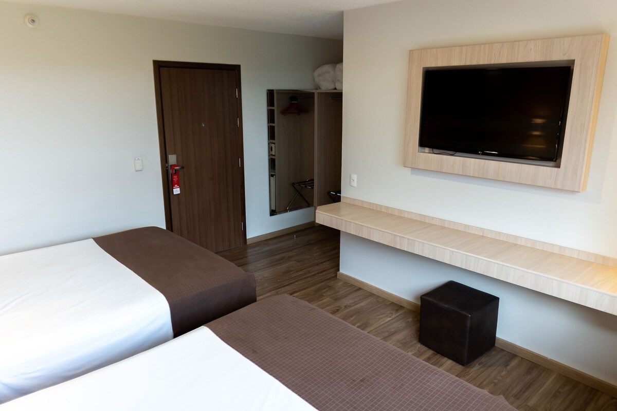 Habitación de hotel con dos camas, televisión montada en la pared, armario y un taburete pequeño.