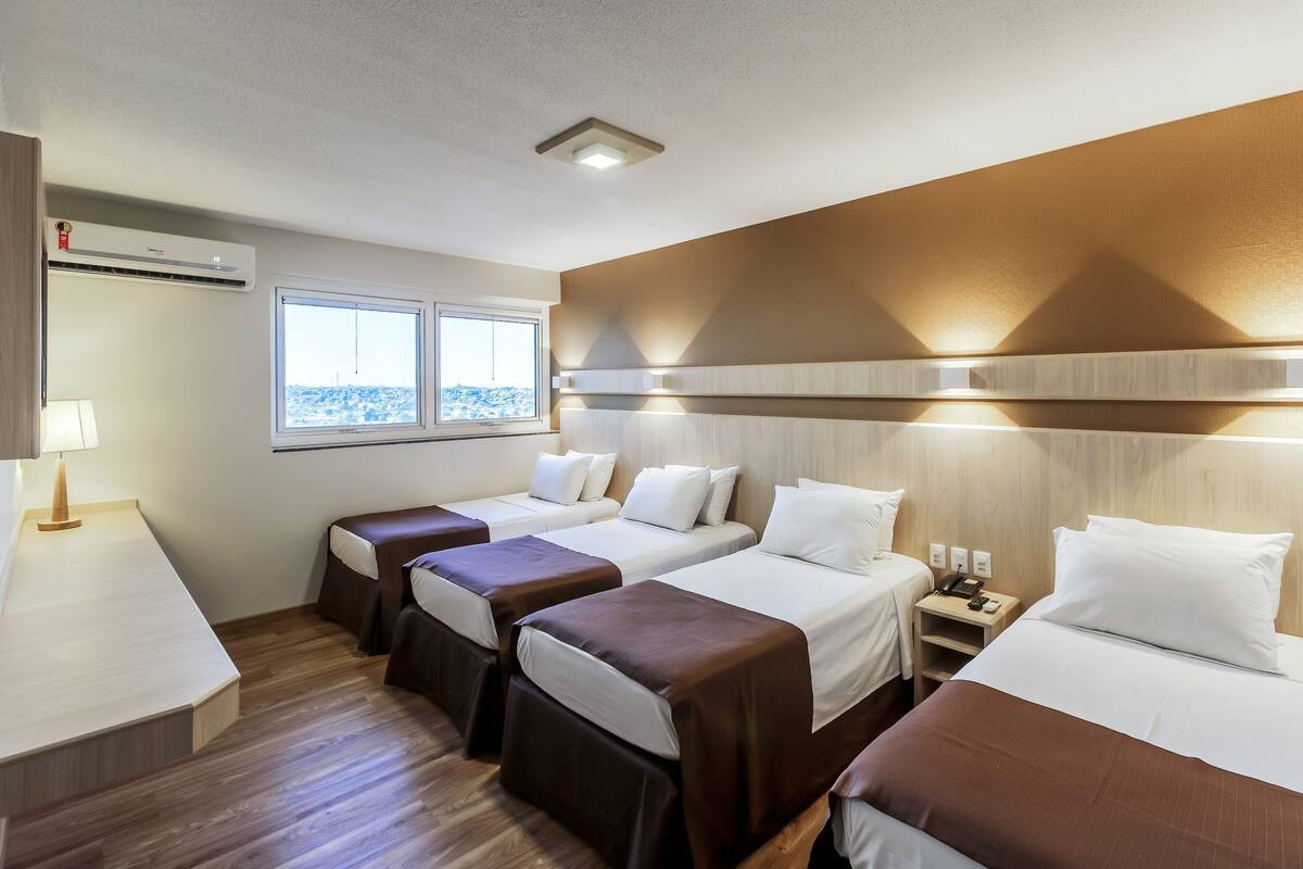Habitación de hotel con tres camas, un escritorio, una lámpara, un ventanal y un equipo de aire acondicionado.