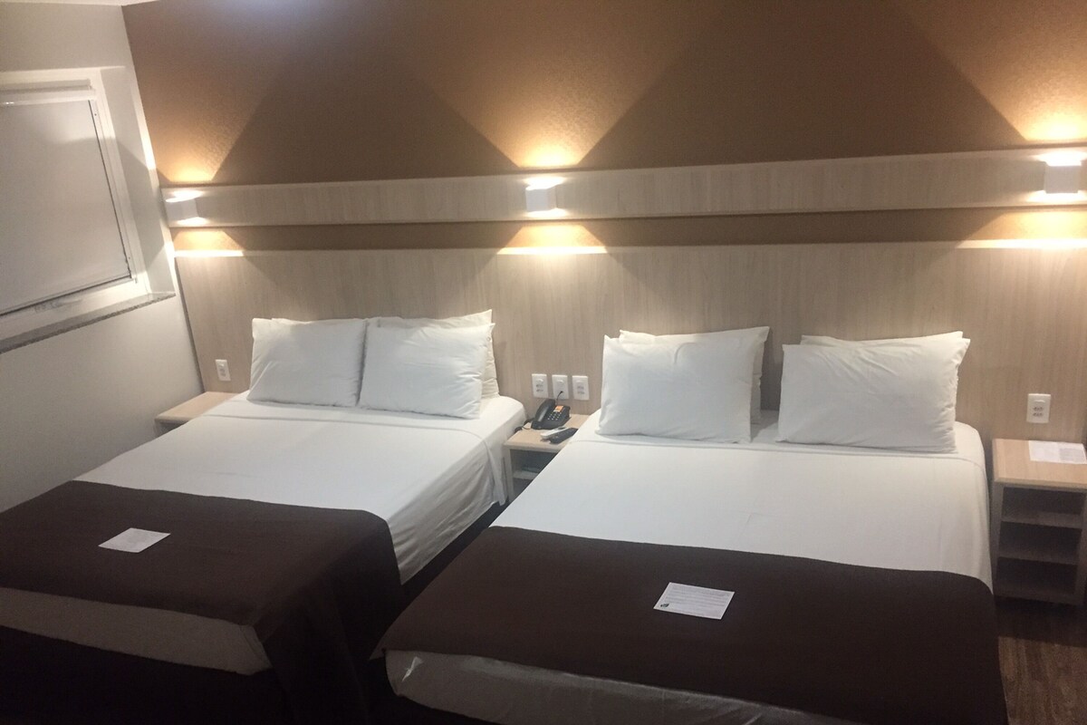 Dos camas individuales con ropa blanca y cabeceros marrones, mesitas de noche con lámparas, y un aplique de pared.