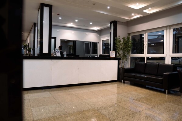 A reception area with a counter, a black leather couch, and a large window. -  Luz Hotel by Castelo Itaipava (Foz do Iguaçu)