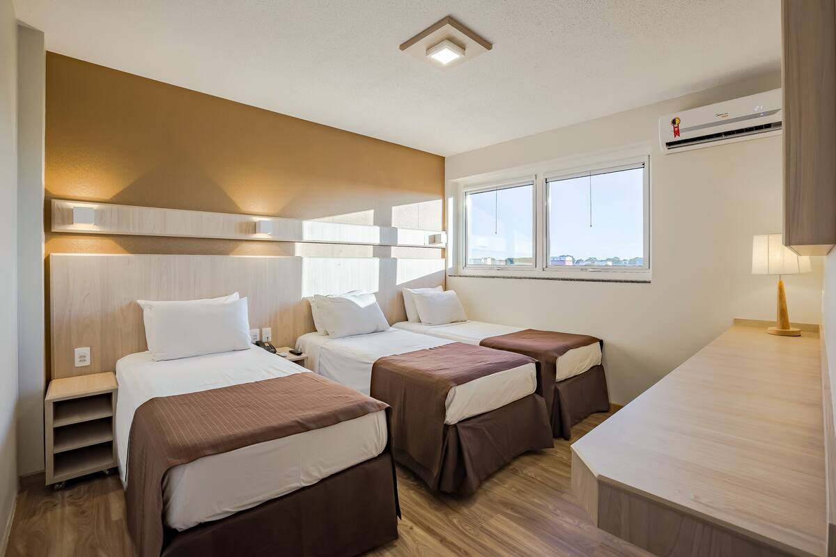 Habitación de hotel con tres camas, un escritorio y ventana con vista a la ciudad.