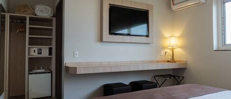 A hotel room with a bed, a TV mounted on the wall, a small desk with a lamp, and a built-in mini-fridge.