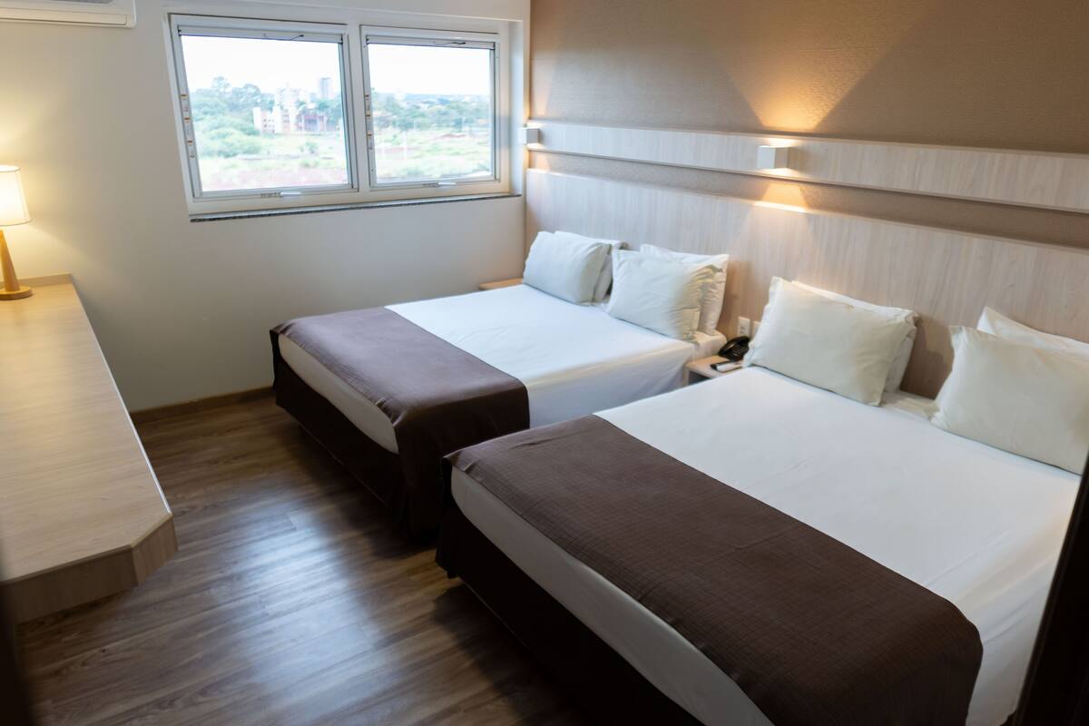 Habitación de hotel con dos camas, una mesita de noche, una lámpara y una ventana que ofrece vistas a zonas verdes y edificios.