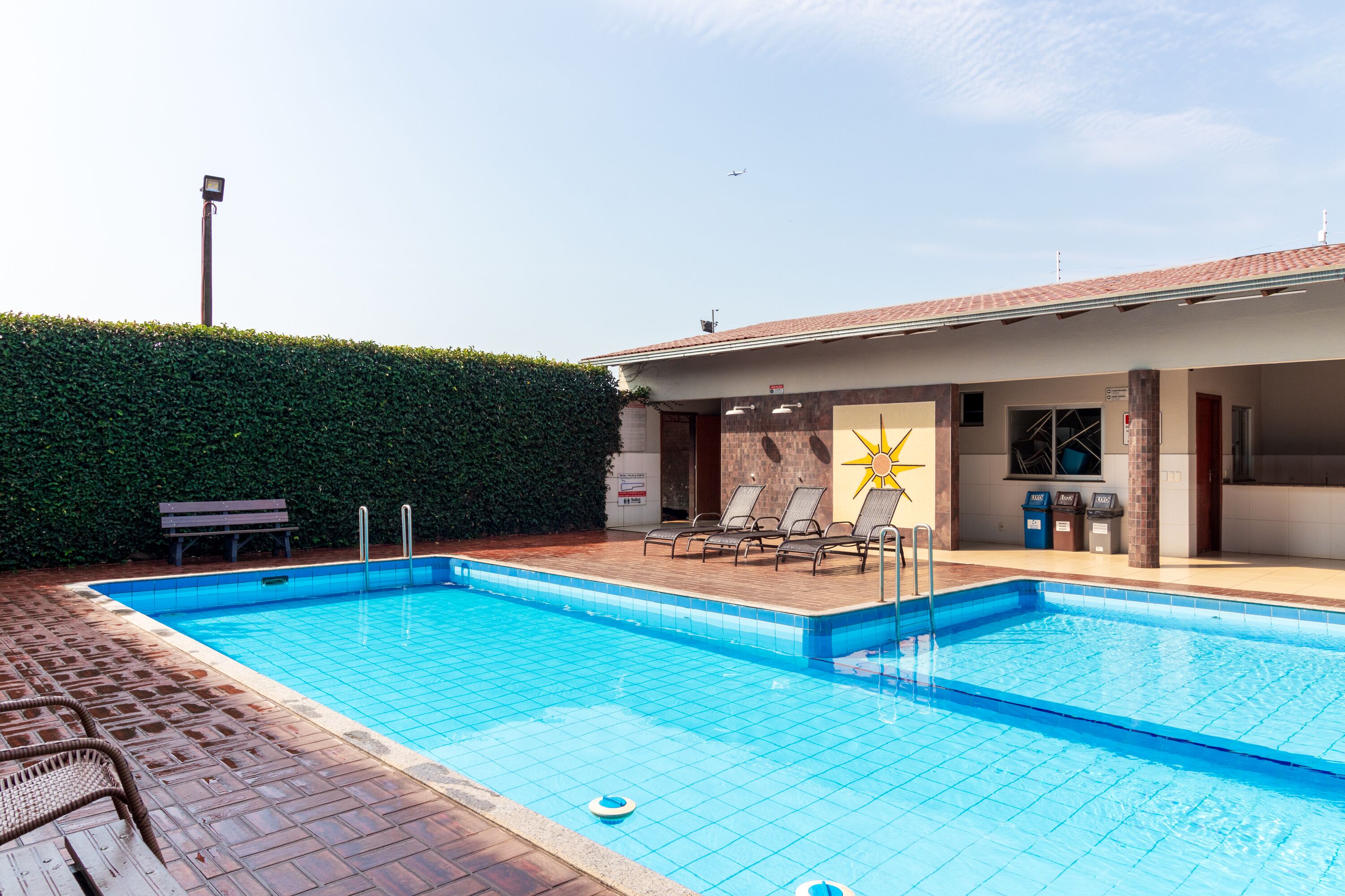 Piscina retangular com borda azulejada, rodeada por um pátio de tijolos. Há espreguiçadeiras e um banco perto da piscina, e um edifício com telhado de azulejos ao fundo.