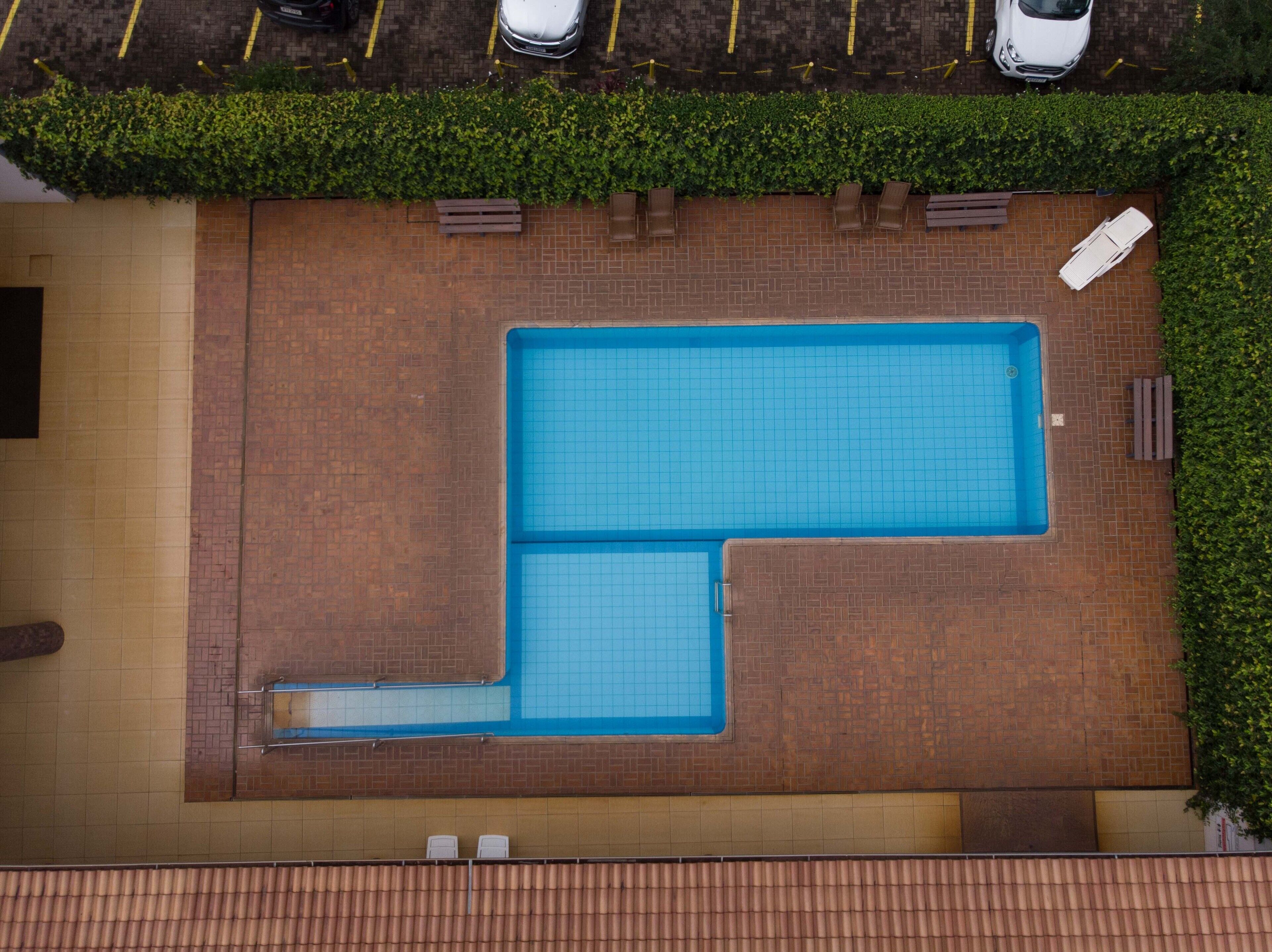 An aerial view of a rectangular swimming pool with a smaller adjacent pool, surrounded by a brick wall and a green hedge.