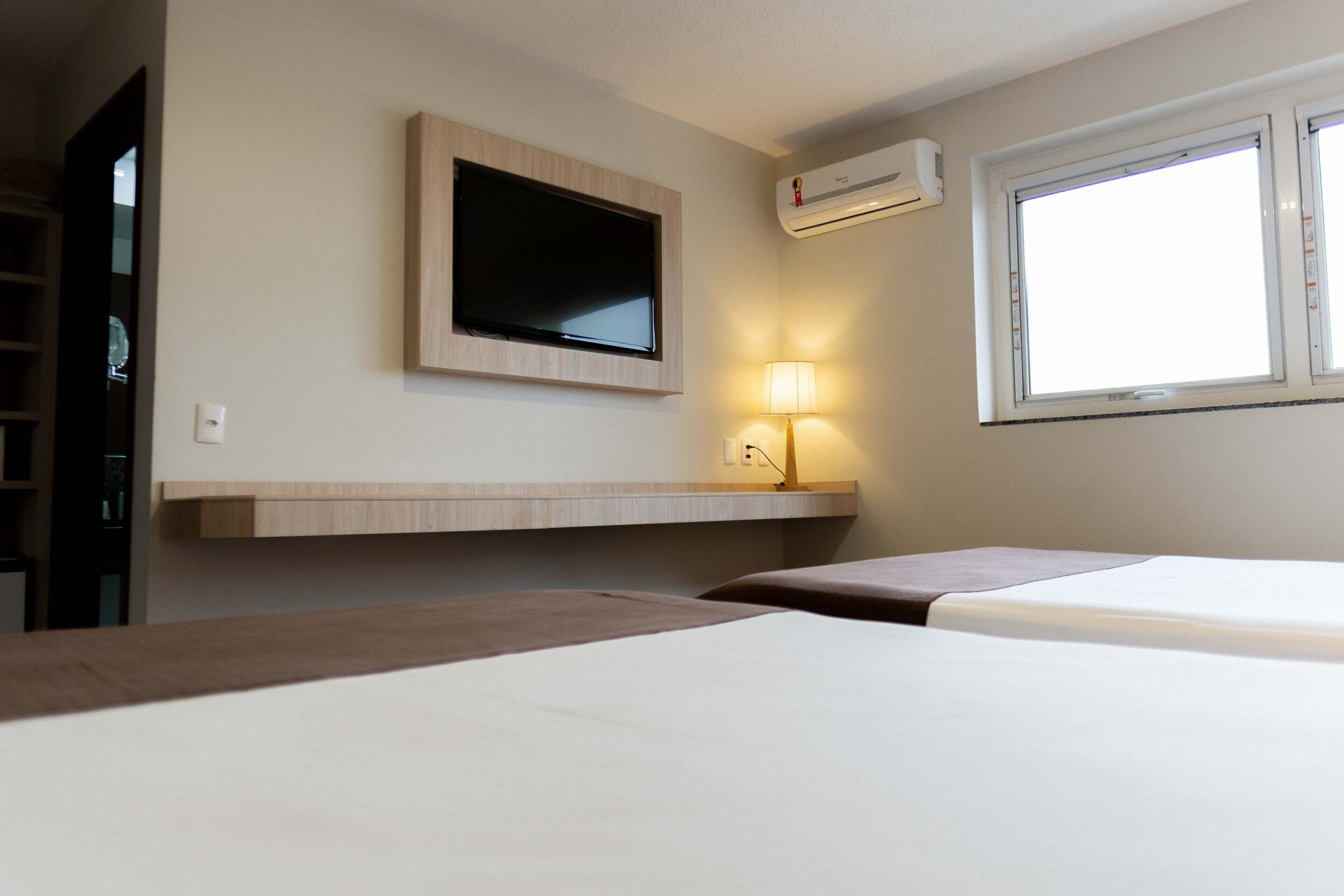 A hotel room with two beds, a mounted TV, an air conditioning unit, and a window with natural light.