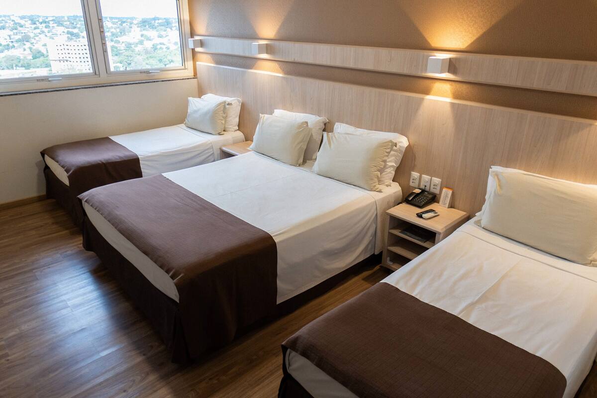 Habitación de hotel con tres camas, cada una con cabecero marrón y ropa de cama blanca. Hay una mesita de noche con un teléfono y una lámpara.