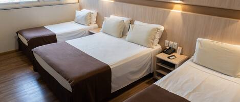 Quarto de hotel com três camas, cada uma com cabeceira marrom e roupa de cama branca. Há uma mesinha de cabeceira com telefone e luminária.