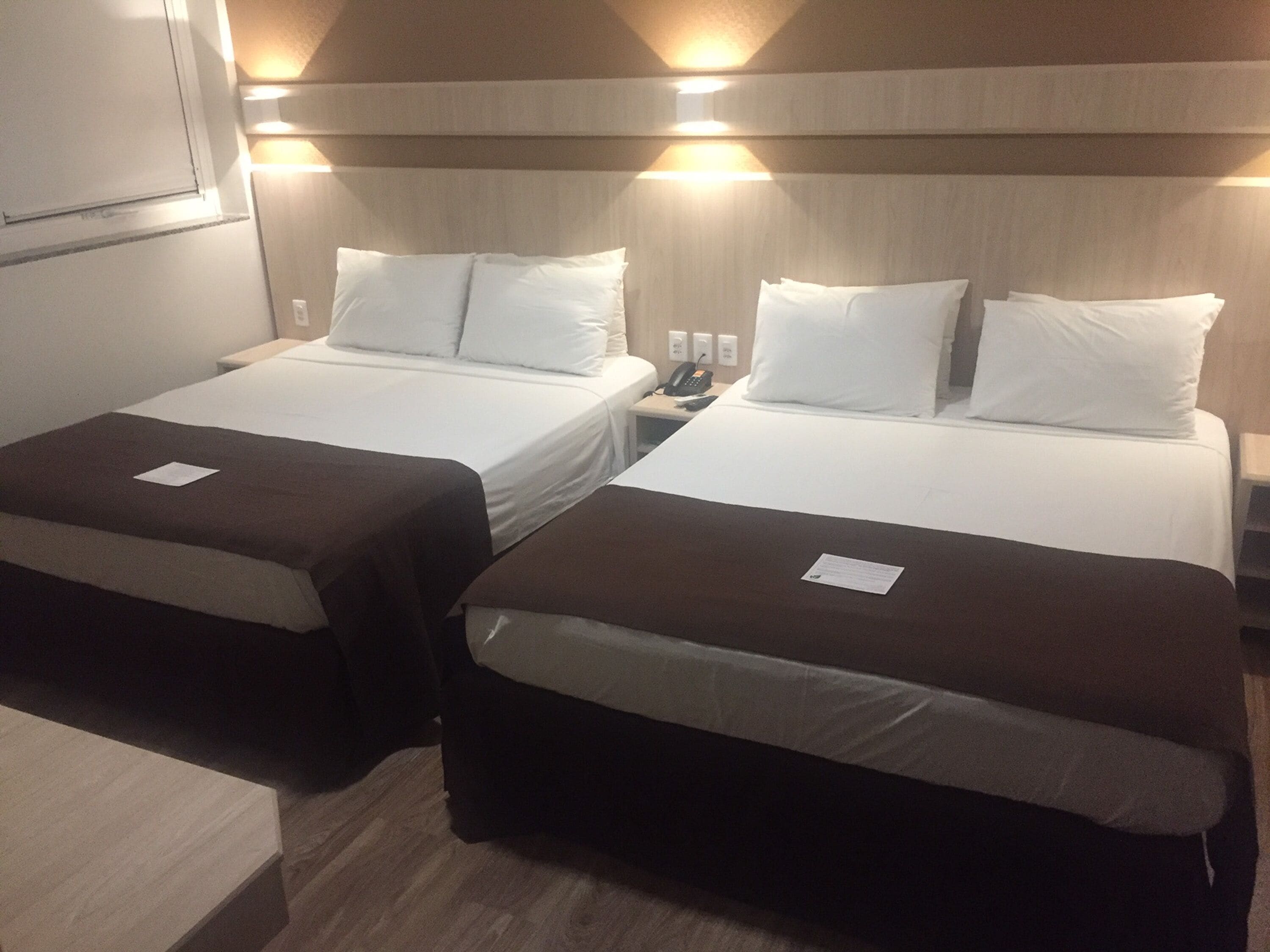 A hotel room with two single beds, each with a brown bedspread and white pillows. There is a bedside table with a lamp and a phone on it. The room has a wooden headboard and wall-mounted lights.