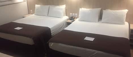 Habitación de hotel con dos camas individuales, cada una con cubrecama marrón y almohadas blancas. Hay una mesita de noche con una lámpara y un teléfono encima. La habitación tiene un cabecero de madera y luces fijadas a la pared.