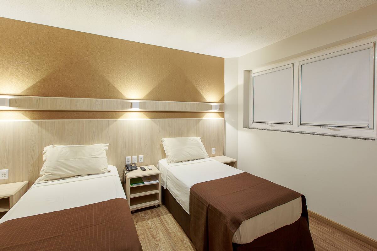Habitación de hotel con dos camas, una mesita de noche con lámpara, una pequeña mesa y una ventana con persianas.