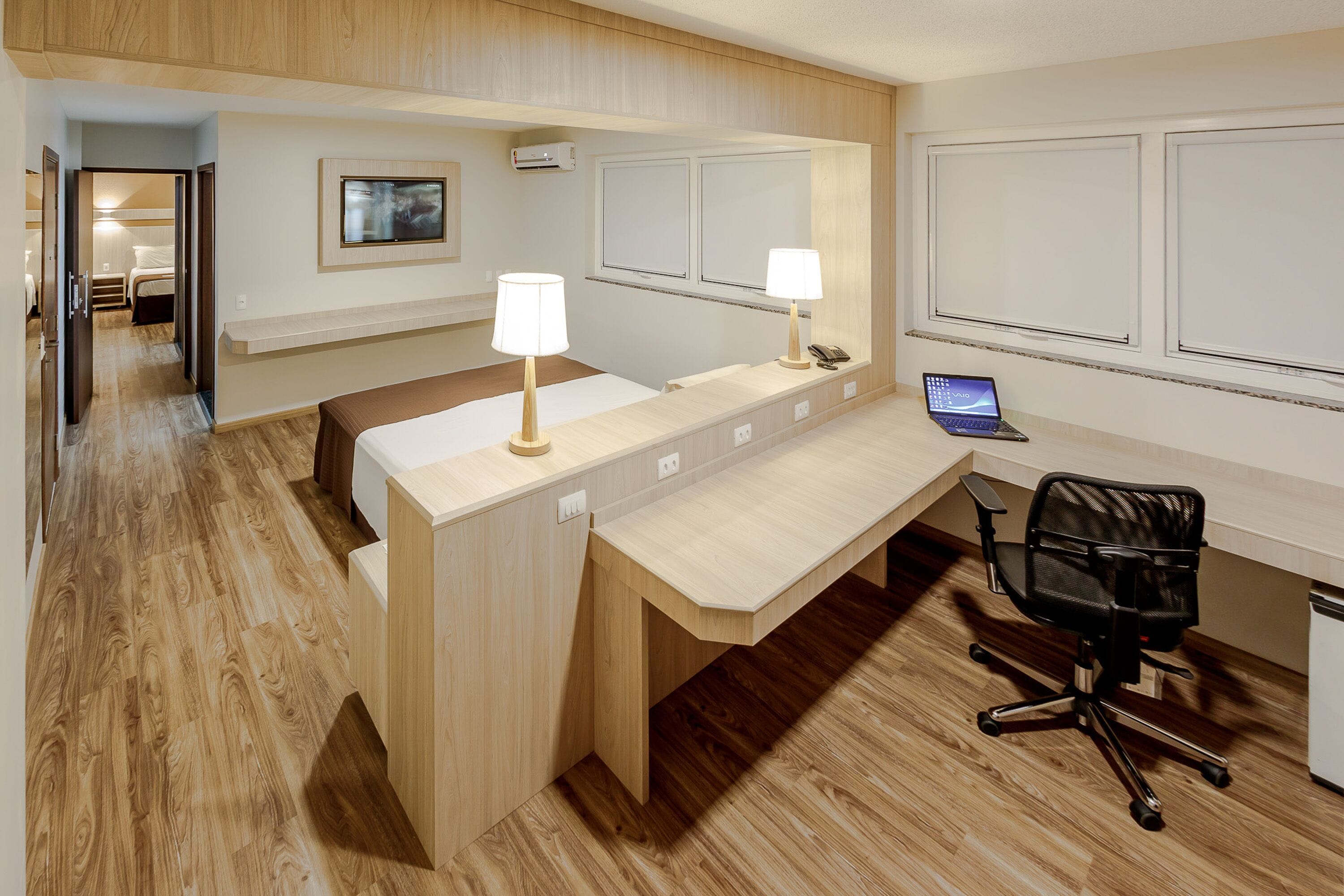 Een hotelkamer met een houten bureau, twee lampen, een laptop en een zwarte kantoorstoel.