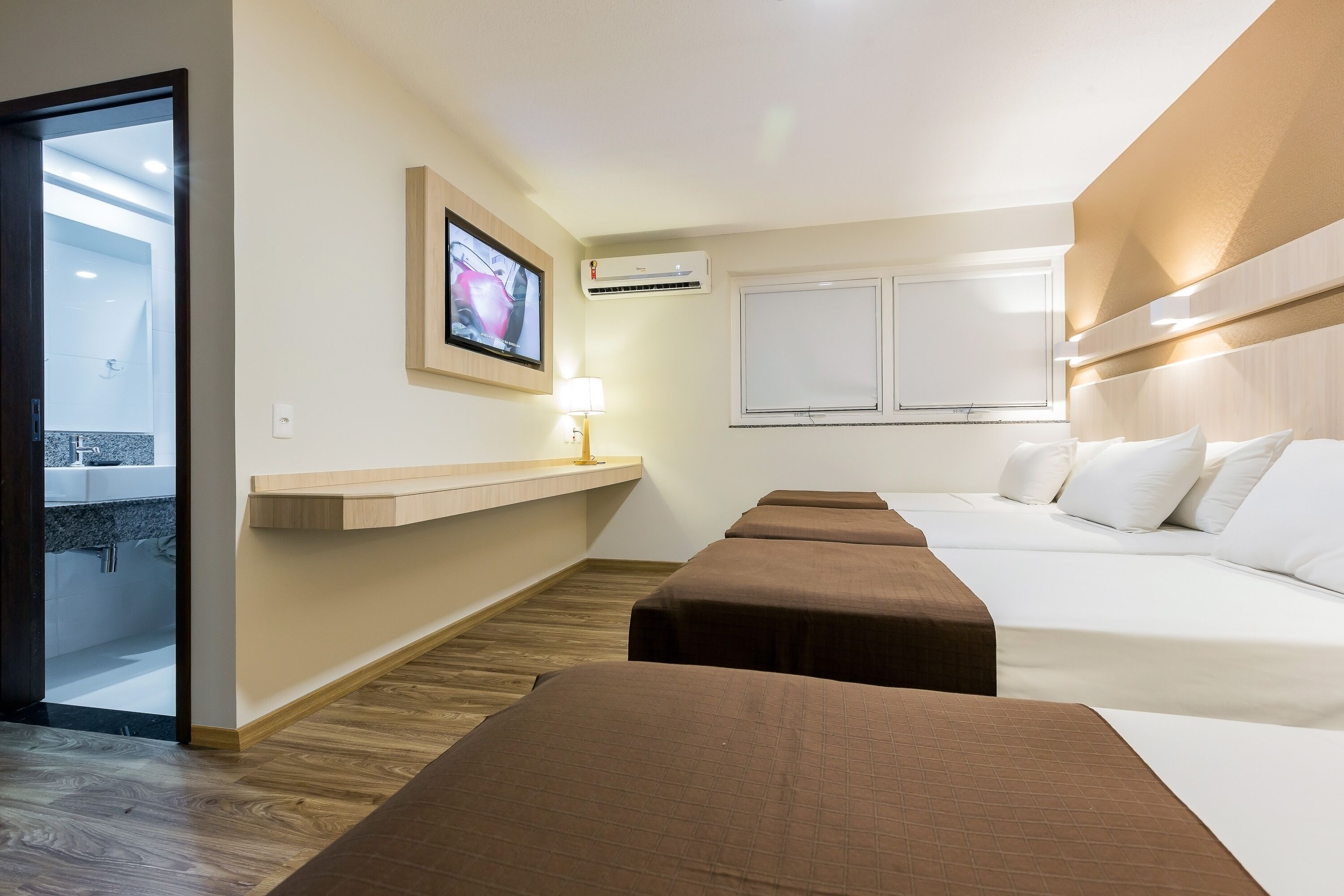 A hotel room with a bed, bedside tables, a TV, an air conditioner, and a window.