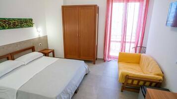 Un dormitorio con cama, armario, una mesita, sofá y ventana con cortinas.