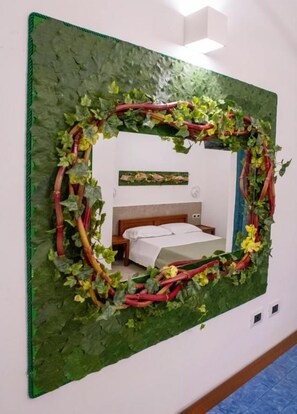 Ein Spiegel mit einem Rahmen aus grünen Blättern, ein Bett mit weißer Bettwäsche und ein Holz-Kopfteil.