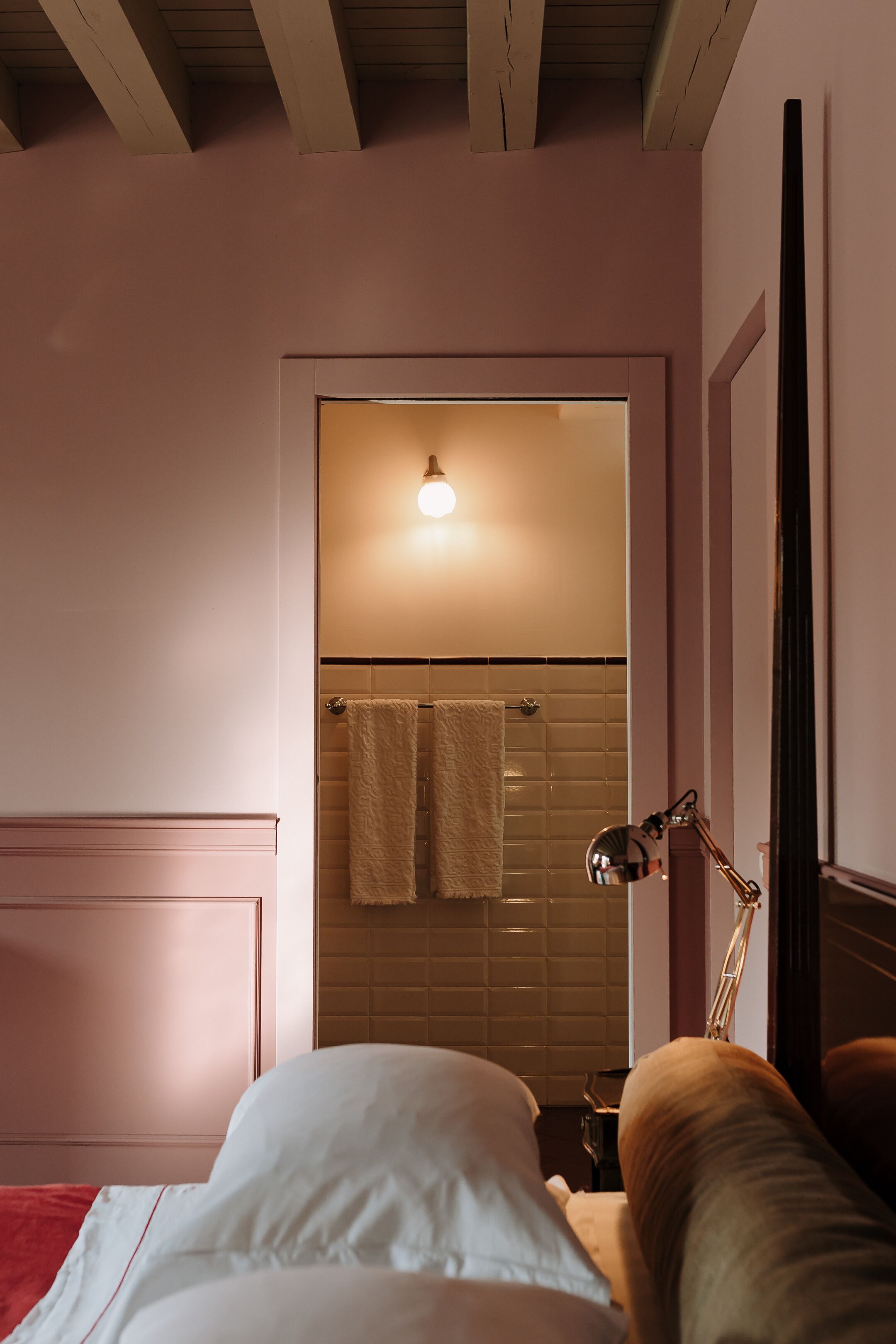 A bedroom with a bed, a nightstand, a lamp, and a door leading to a bathroom with a towel hanging on the wall.