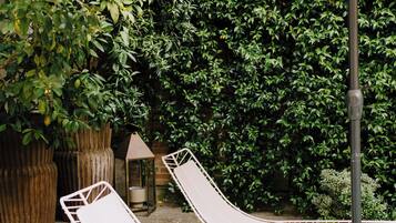 Un jardín con dos sillones de metal blanco bajo un paraguas grande, rodeado de abundante follaje verde y una jardinera de madera.