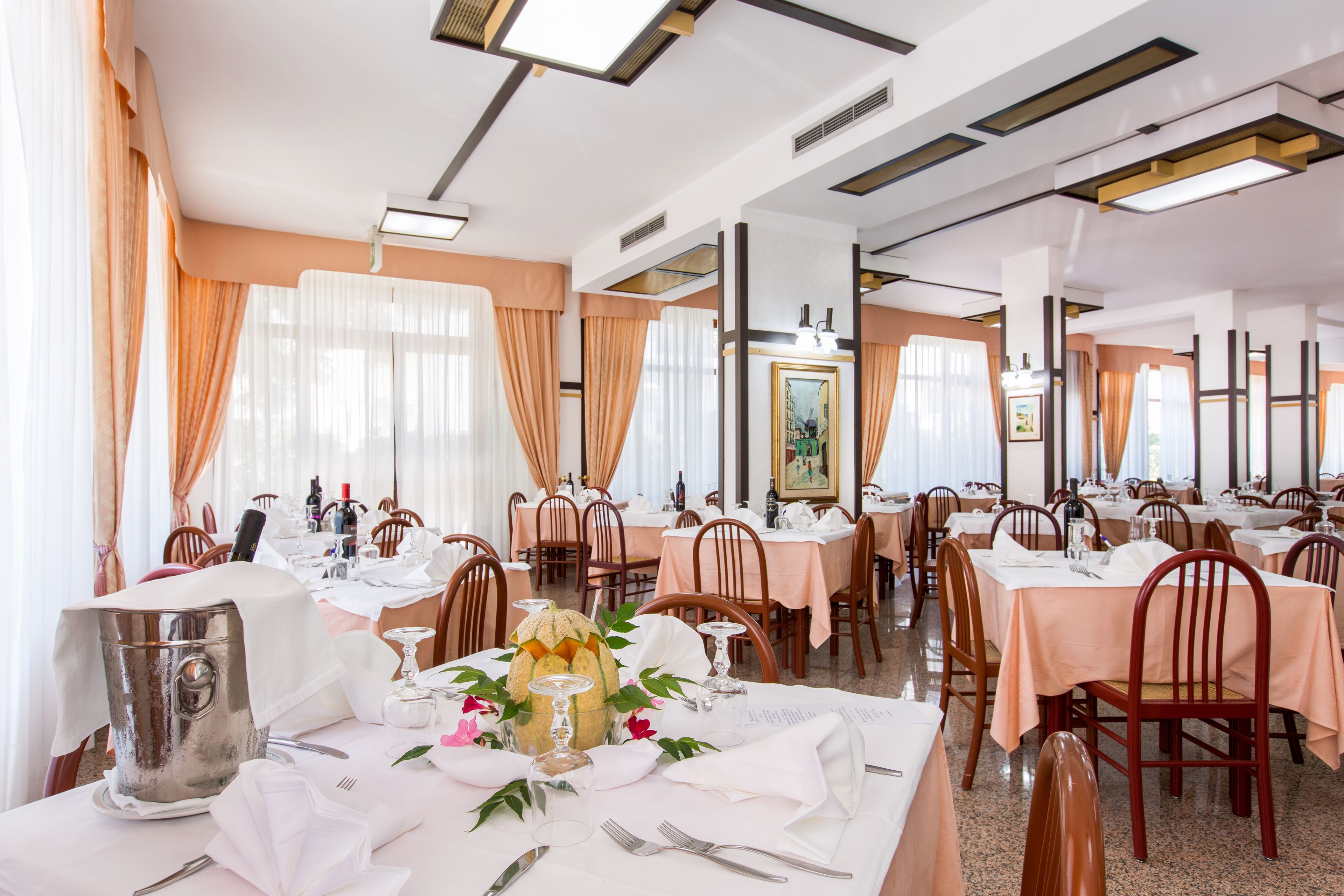 Comedor con mesas dispuestas para una comida, destacando un arreglo floral central y una jarrón de plata.