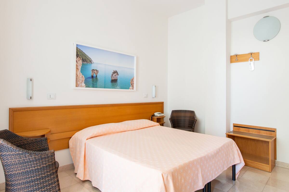 Una habitación de hotel con una cama, una silla, una mesita, una mesita de noche, un espejo y un cuadro de una escena costera.