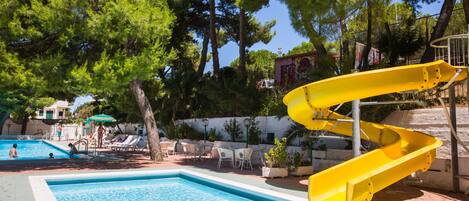 A swimming pool with a yellow water slide, lounge chairs, and a view of trees.