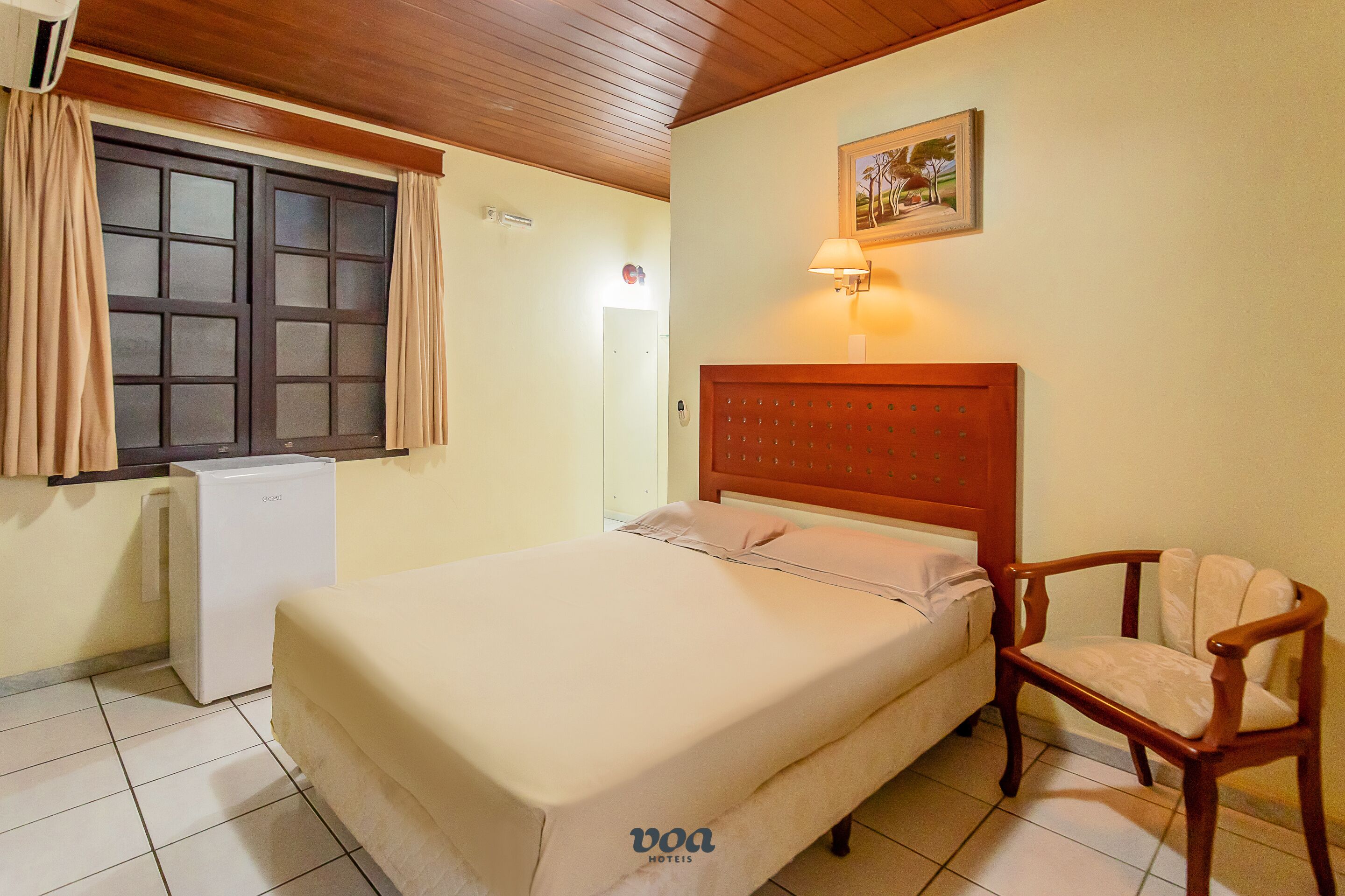 A hotel room with a bed, a chair, a small refrigerator, a window with curtains, and a wall-mounted lamp.