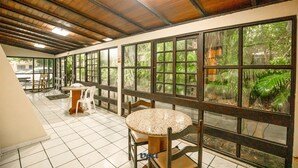 A corridor with a tiled floor, wooden ceiling, and large windows with a view of greenery. There are tables and chairs along the corridor. - VOA Samuka Hotel (Florianópolis)