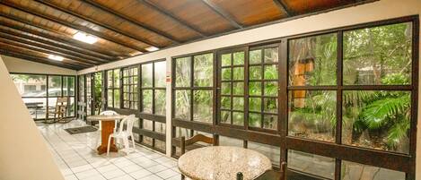 A corridor with a tiled floor, wooden ceiling, and large windows with a view of greenery. There are tables and chairs along the corridor.