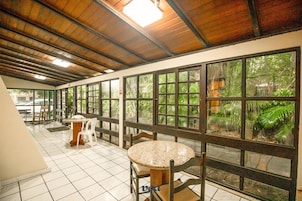 Um corredor com chão de azulejos, teto de madeira e amplas janelas com vista para o verde. Há mesas e cadeiras ao longo do corredor.