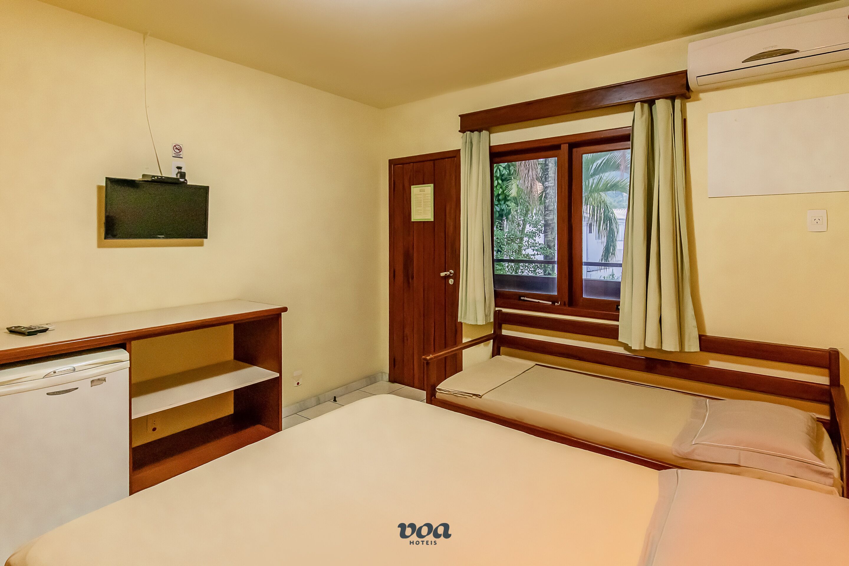 Ein Hotelzimmer mit Bett, Schreibtisch, kleinem Kühlschrank, einem Fenster mit Vorhängen und Blick auf Grünfläche.