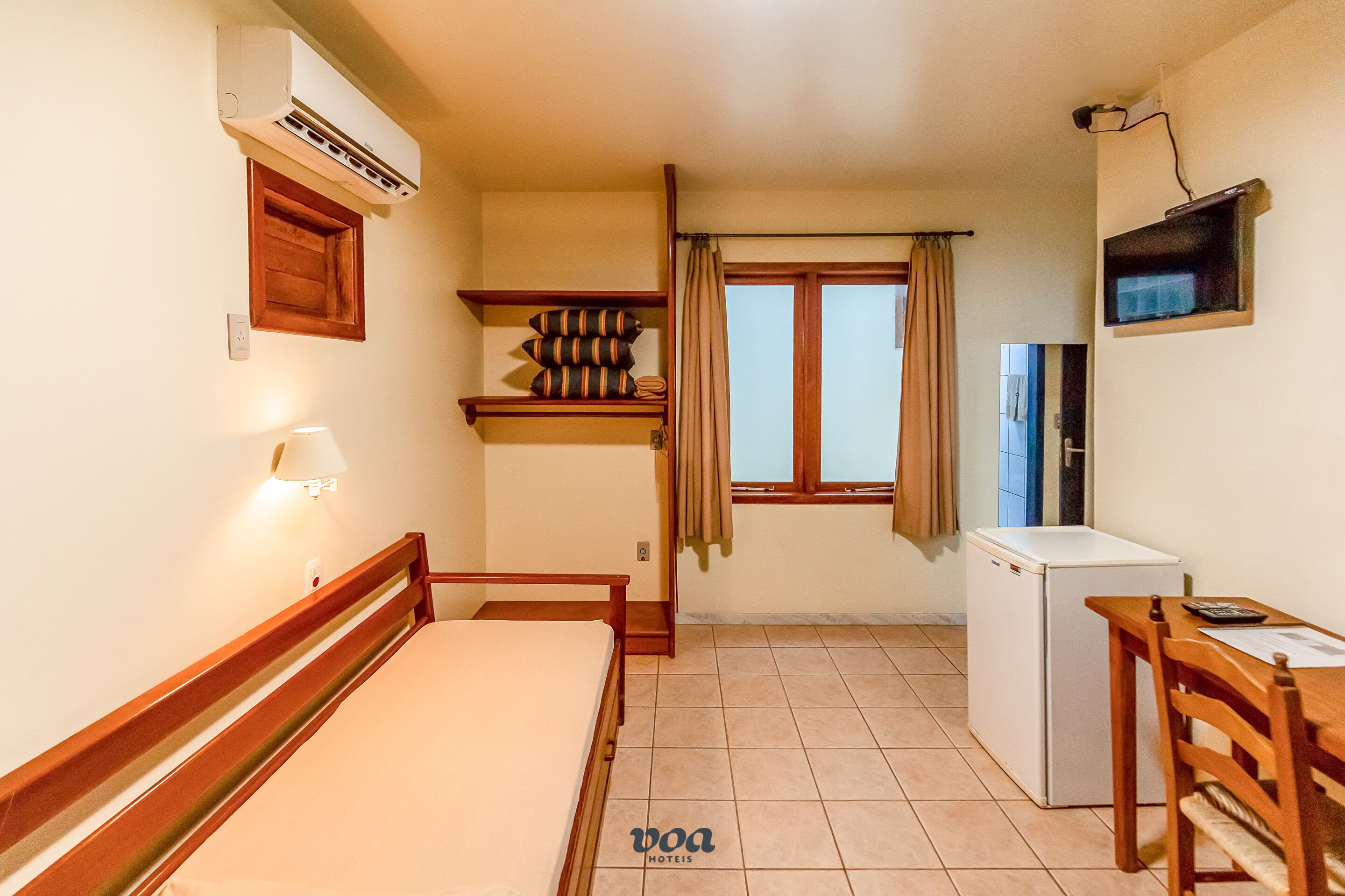 Una camera d'albergo compatta con un letto, una piccola scrivania, un frigorifero, una finestra con tende, un condizionatore d'aria a parete e uno scaffale con cuscini.