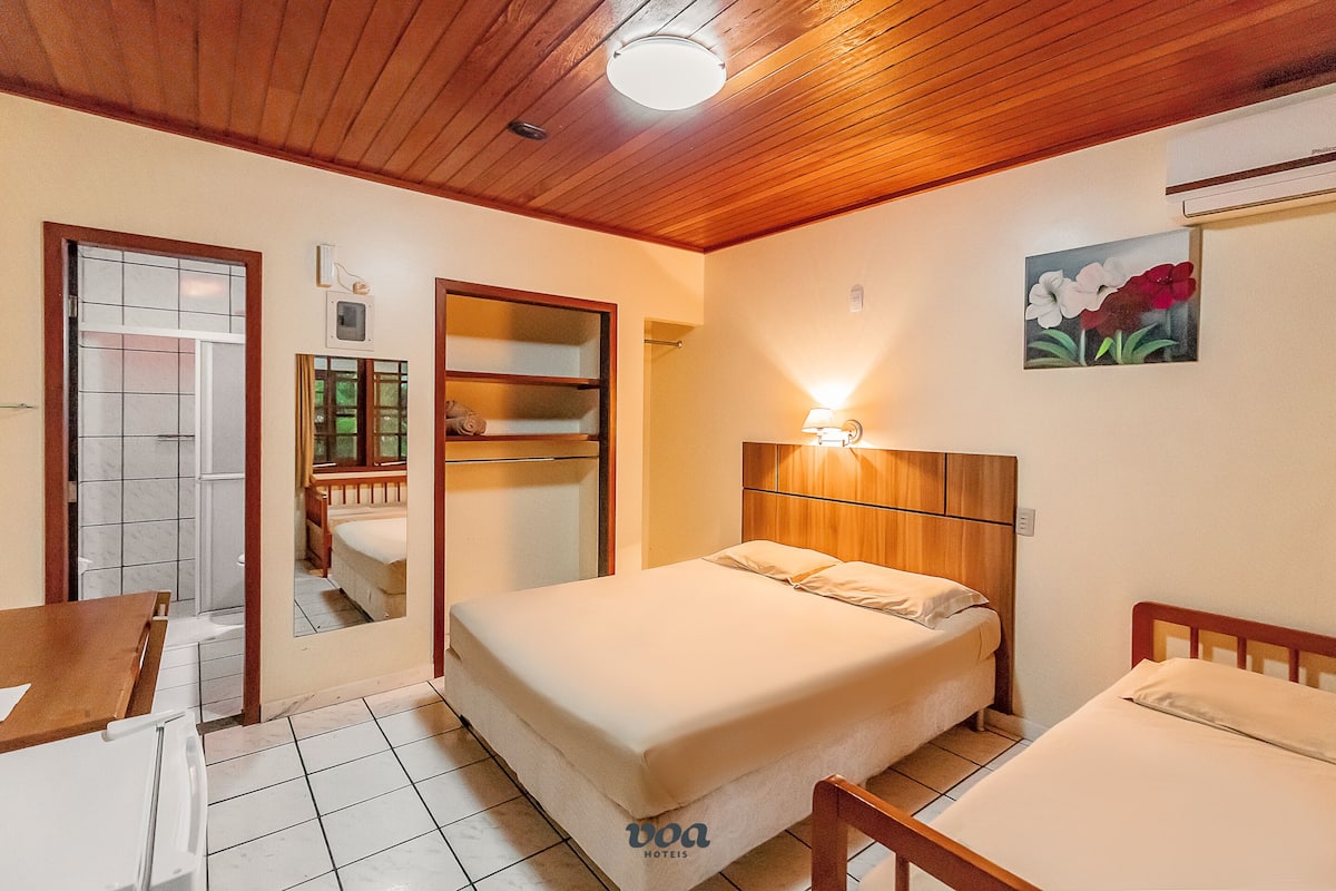 A hotel room with a wooden ceiling, a bed with a wooden headboard, a bedside table, a chair, a small table, a painting on the wall, and a window with a view of greenery.