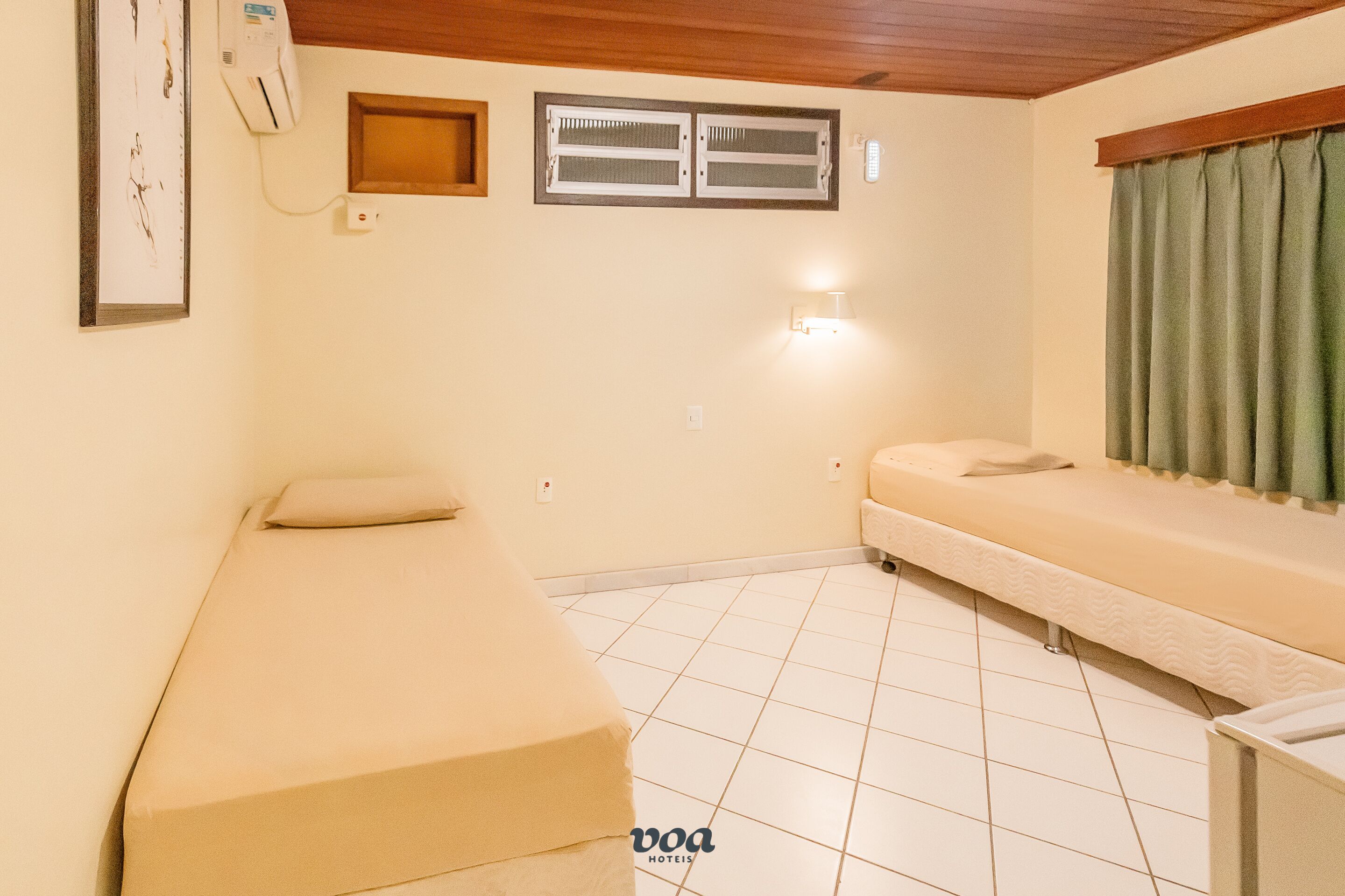 A small, single-room hotel room with a bed, a bench, a window, and a wall-mounted air conditioner.
