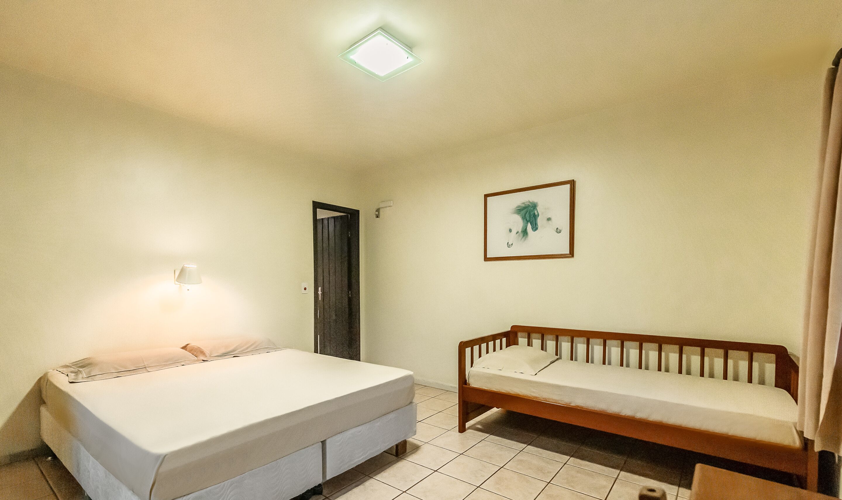 Una habitación pequeña y limpia con una cama, un banco de madera, un cuadro en la pared y una puerta.
